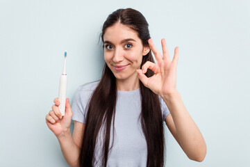 Young caucasian woman holding a electric toothbrush isolated on blue background cheerful and confident showing ok gesture.