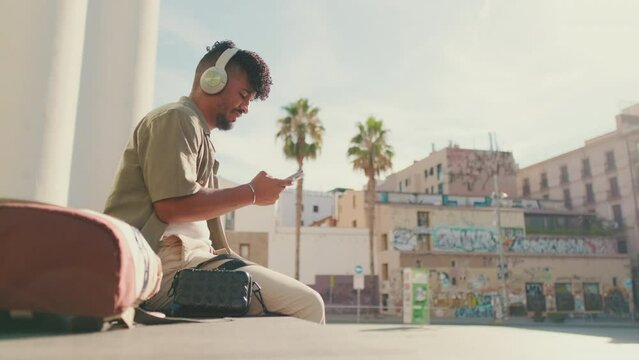 Young male student dressed in an olive color shirt sits outside next to the university, listens to music on headphones, selects tracks on his phone