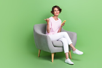 Fototapeta na wymiar Full body photo of nice young lady hold gadget point empty space sit chair wear stylish striped outfit isolated on green color background