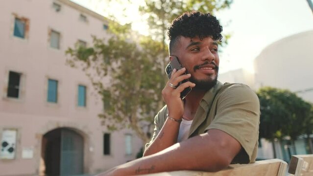 Young man with beard wearing an olive colored shirt with headphones talking on the phone while sitting on bench on modern buildings background