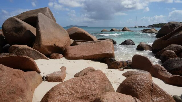 The waves of the turquoise ocean beat against granite boulders and splash on the sandy beach. A white yacht is visible in the distance. Blue sky with clouds. Seychelles. Praslin Island. Anse Lazio bea