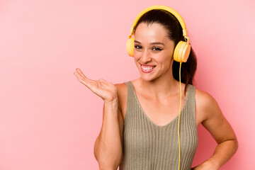 Young caucasian woman listening to music isolated on pink background showing a copy space on a palm and holding another hand on waist.