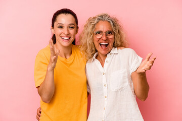 Caucasian mom and daughter isolated on pink background receiving a pleasant surprise, excited and raising hands.