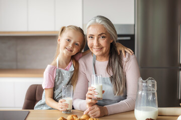 Smiling european old female and little girl in aprons with glasses of milk enjoy croissants in kitchen interior