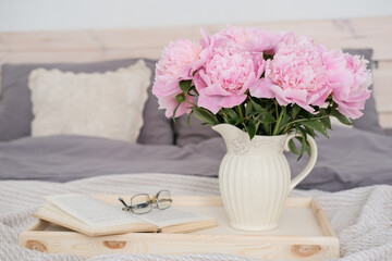 A vase with a bouquet of pink peonies, an open book and glasses on a tray on the bed.