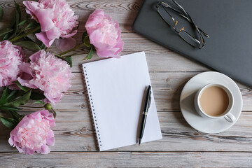 Top view of feminine workspace with laptop, blank notepad, cup of coffee and peonies flowers. Women's work office. Mock up notepad