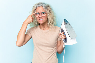 Middle age caucasian woman holding an iron isolated on blue background showing a disappointment...