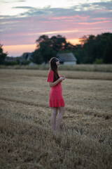 Beautiful pensive young girl in a red dress with long hair walking on a stubble on a hillside at sunset after harvest in the countryside, rural landscape, Poland