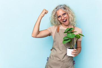 Middle age gardener woman holding a plant isolated on blue background raising fist after a victory,...