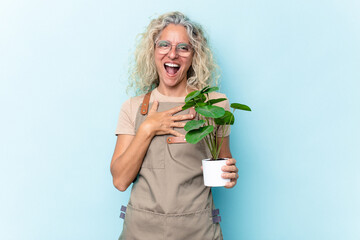 Middle age gardener woman holding a plant isolated on blue background laughs out loudly keeping...