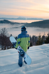 man snowboarder enjoying the view of sunset above the snowed mountains