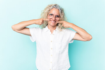 Middle age caucasian woman isolated on blue background smiles, pointing fingers at mouth.