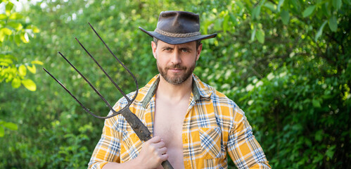 Serious guy holding hayfork. Unshaven guy wearing cowboy hat and plaid shirt outdoors. Guy farmer