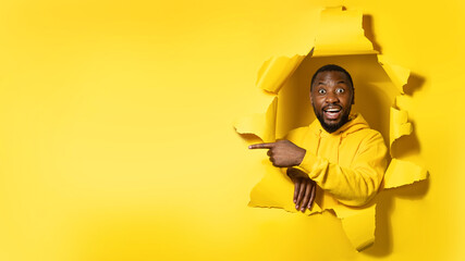 Cheerful black man pointing aside recommending best offer, peeking out through torn yellow paper hole, free space
