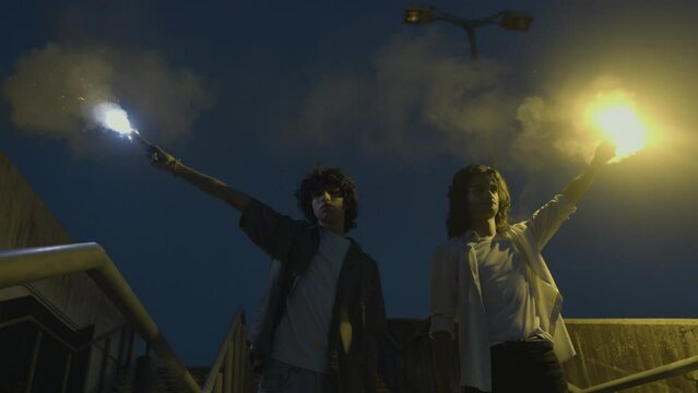 Kids holding flare torches, standing in urban area at night time, low angle view medium shot. Teenagers protest, rebellion demonstration, youth concept