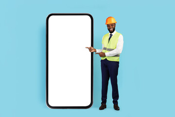 Construction Offer. Smiling Black Male Engineer Pointing At Huge Blank Smartphone