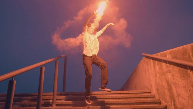 Teenager running down urban stairs with burning signal flare at night, jumping, waving torch, slow motion tracking shot. Youth, rebellion expressive demonstration, teen freedom concept