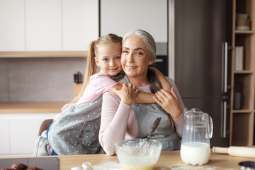 Smiling little girl hugs old woman in apron making dough in kitchen interior