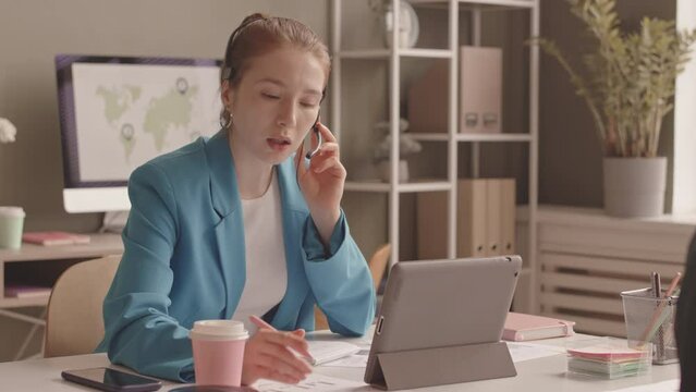 Waist up slowmo of pretty young Caucasian red haired woman working at call center in office, speaking to client via headset and using digital tablet