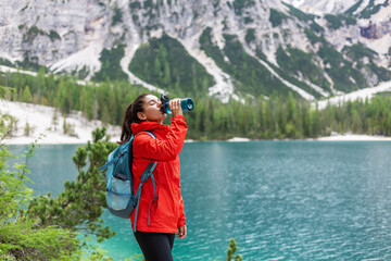 Hydration while on track. Attractive woman hiker in red raincoat drinking water near a lake and...