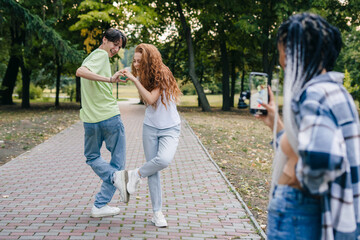 Obraz premium Cheerful beautiful girl standing in the park with a camera and smiling while recording a video of her two friends dancing while showing the heart sign