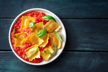 Ravioli with tomato sauce and fresh basil leaves on a plate, Italian pasta dish, shot from the top on a dark wooden background with copy space