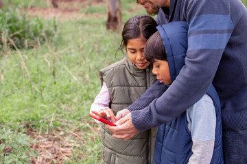 Boy wearing a blue coat and a girl wearing a green coat with a man looking at a red mobile phone