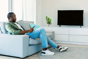 Rest and relax. Black man sitting on the sofa in living room, watching television show, looking at...