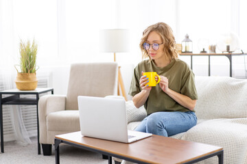 Woman glasses using laptop computer while sitting sofa with big window on background at home interior