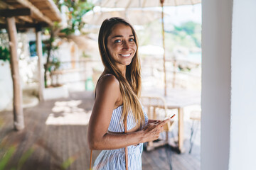 Cheerful young lady smiling at camera while using smartphone in summer cafe