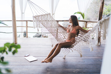 Fit young female traveler relaxing in hammock on wooden veranda at seaside - obrazy, fototapety, plakaty