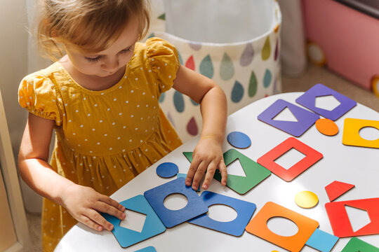 A little girl playing with wooden shape sorter toy on the table in playroom. Educational boards for Color and Shapes sorting for toddler. Learning through play. Developing Montessori activities.