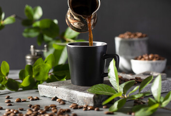 Coffee is poured into a black mug from a metal cup on a dark background.