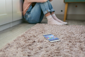 Mobile phone lying on the floor with unrecognizable sad little girl sitting behind. Selective focus...