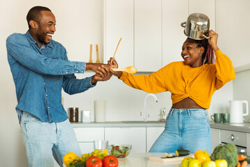 Domestic fun. Playful black couple fighting in kitchen, using spatulas as a weapons, fooling...