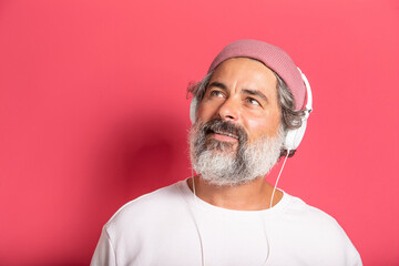 Cheerful grey-haired bearded man in casual pink hat and pullover posing isolated on fuchsia wall background studio portrait. People lifestyle concept. Listen to music with headphones, sing songs