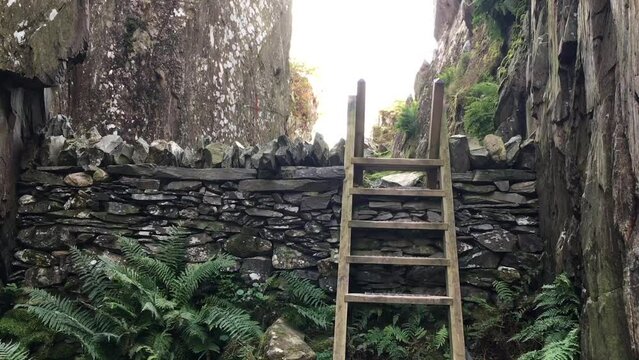 A wooden ladder stile over a dry stone wall, leading into a narrow rock ravine with mountains in the background. Llyn Idwal, Snowdonia National Park, Wales.