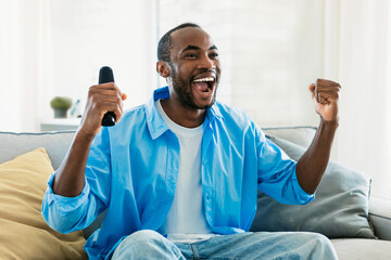 Overjoyed black man watching sport on TV at home, celebrating success, raising fists up and screaming, copy space