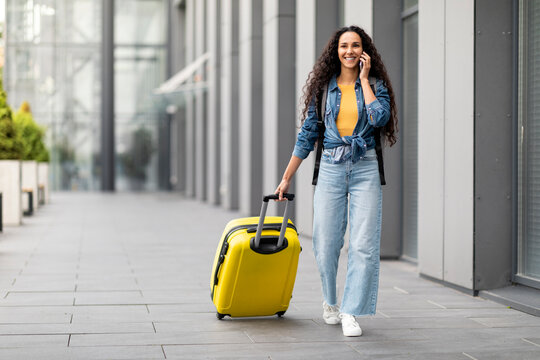 Smiling young woman traveller walking by airport, having phone conversation