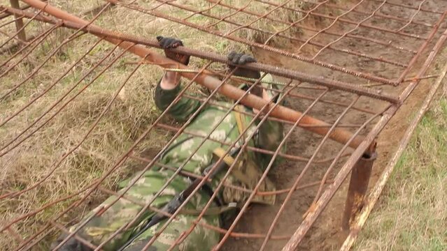 Military soldiers crawl under a net on their backs wearing a gas mask and carrying weapons during an obstacle course at a training camp