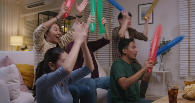 Group of young adult friend man and woman asia people sit at sofa couch joy party fun game FIFA world cup live TV at home chanting hand raised sign jump mad happy win exult face.