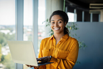 Portrait of positive black female entrepreneur standing near window and using laptop computer, free...