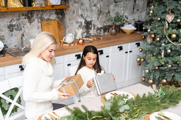 Beautiful mother and daughter have fun, hug and kiss at home near the Christmas tree in a white interior. Family happiness, holiday, joy, vacation, games with a woman. New Year's preparations