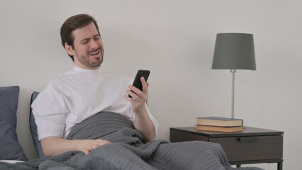 Young Man Reacting to Loss on Smartphone in Bed