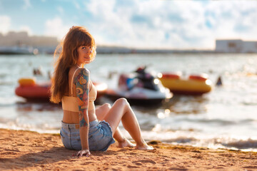 Relax at resort. Young caucasian smiling tanned woman with tattoos poses sitting at the sand beach....