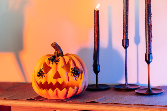 Halloween pumpkin Jack-o-lantern and black candles standing on modern wooden cabinet in orange and blue light. Seasonal living room interior decoration for holidays and festive party