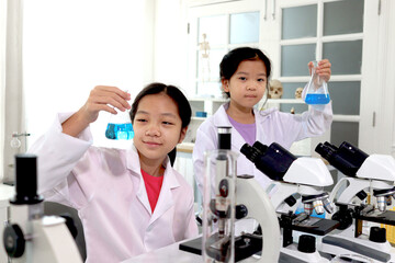 Fototapeta premium Two adorable pretty schoolgirls in lab coat doing simple science experiments, young Asian kid scientist having fun in chemistry laboratory class, happy smiling girl showing chemical flask, little chil