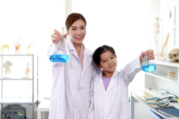 Portrait of beautiful Asian science teacher and adorable schoolgirl in lab coat standing and showing blue chemical flasks to camera in laboratory. Smiling young scientist with kid have fun together an