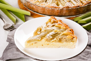 Piece of homemade rhubarb pie with almond frangipane cream and rhubarb stalks on white table. Delicious home baked pastry. High angle