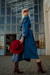 Fashionable confident blonde woman wearing trendy sunglasses, blue midi trench coat, belt, leather...